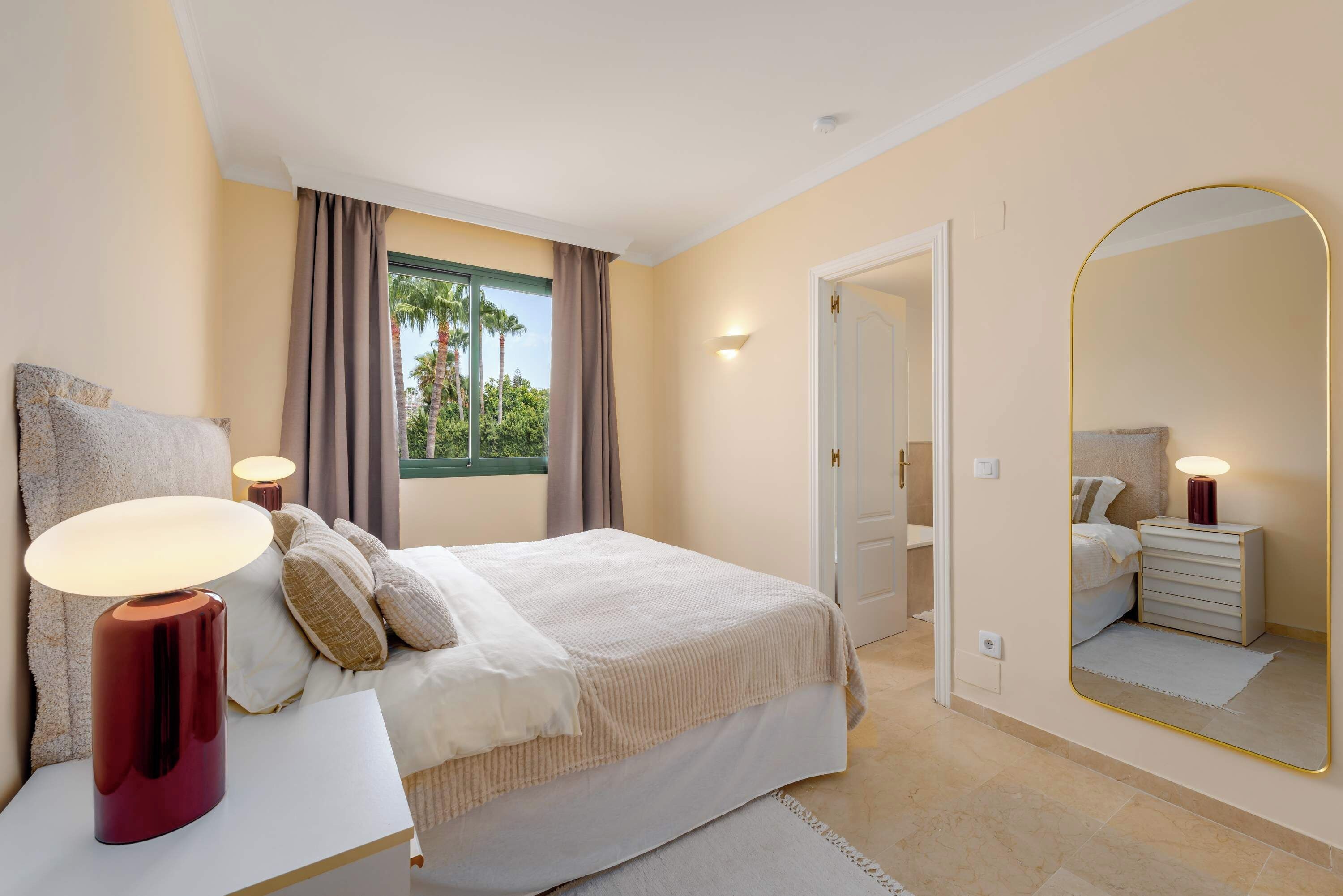 A bedroom with a bed, bedside lamps, a large mirror, and a view of palm trees through the window.