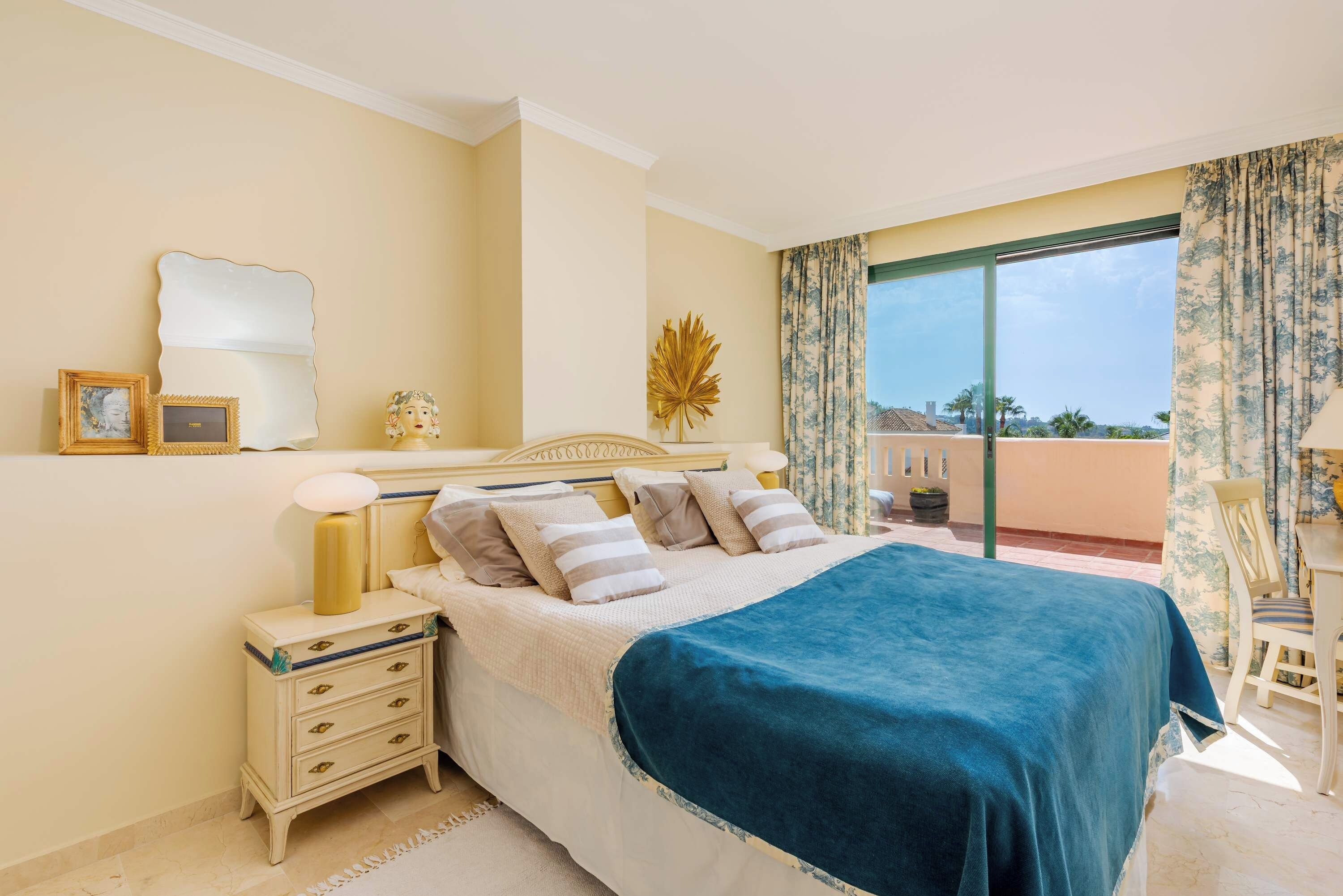 A bedroom with a bed, bedside table, lamp, and a view of the outdoors.