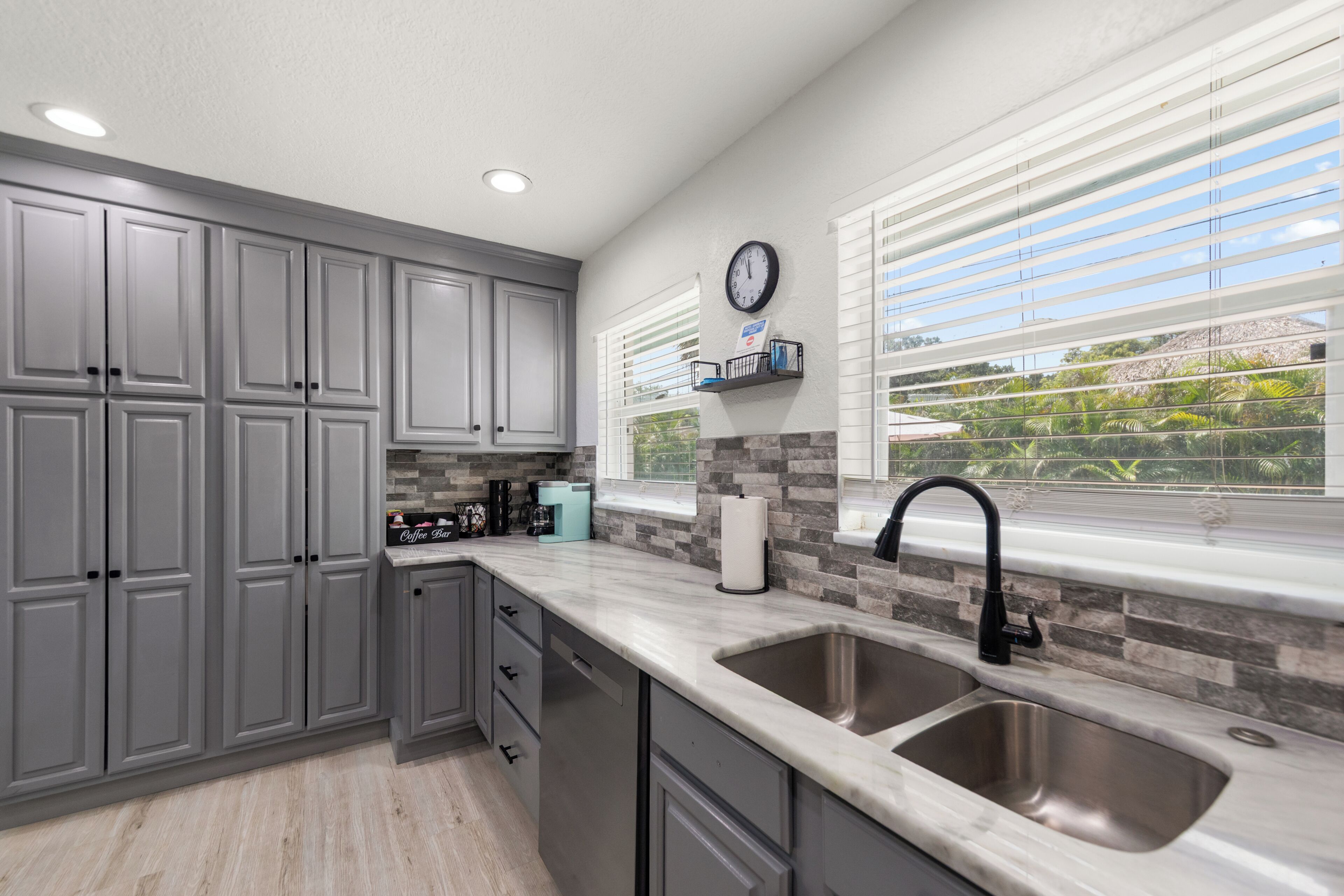 A modern kitchen with grey cabinets, a double sink, a coffee station, and a view of greenery through the window.