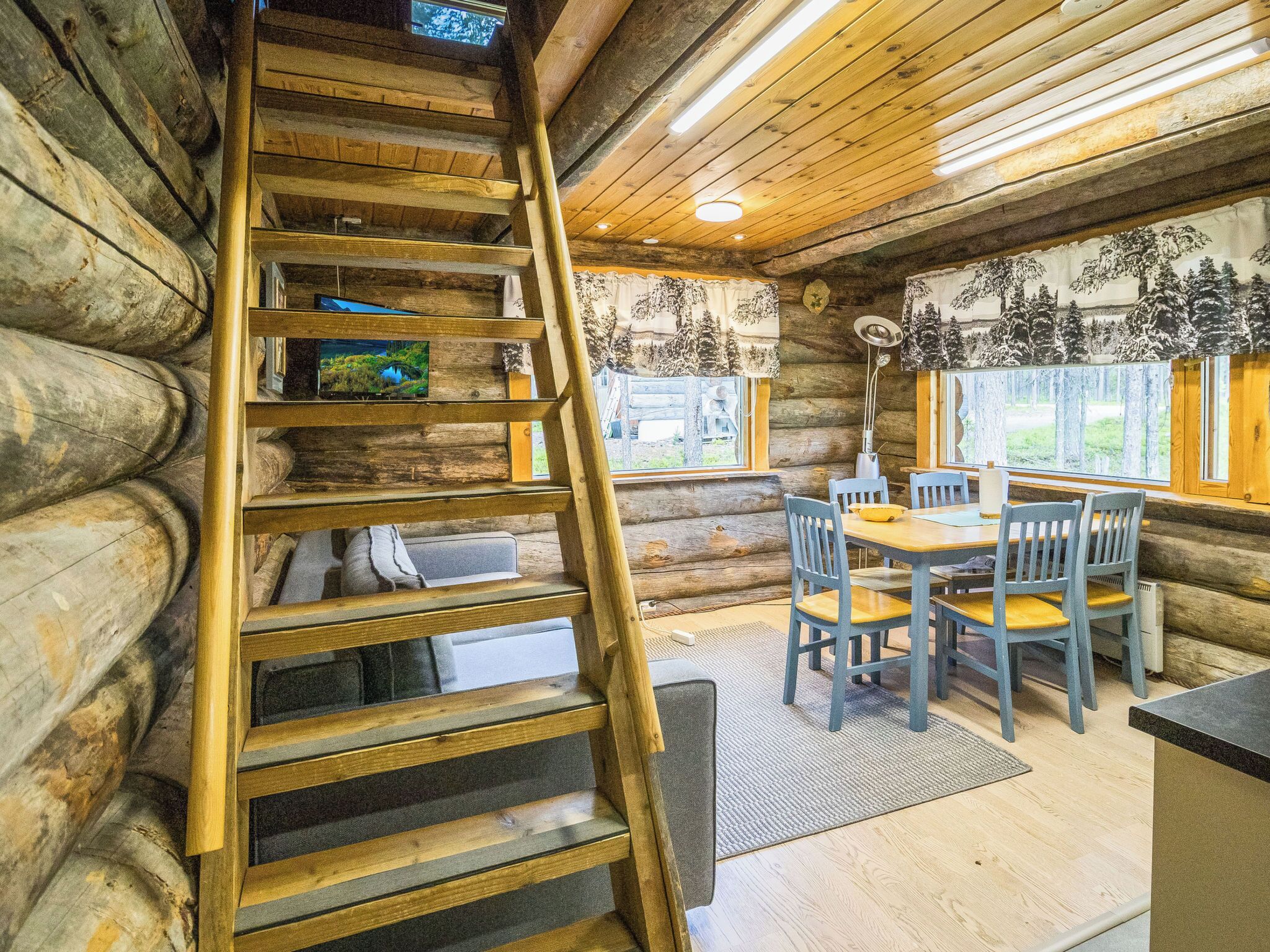 L’intérieur d’une cabane en rondins, avec un escalier en bois, un coin repas avec des chaises bleues et une vue sur l’extérieur par les fenêtres.