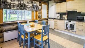 Interior de una cabaña de troncos con comedor, cocina y grandes ventanales con vista a un bosque.