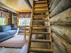 L’intérieur d’une cabane en rondins, avec une échelle en bois, un canapé et une fenêtre donnant sur une forêt.