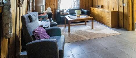 Uma sala de estar aconchegante com sofá, poltrona e mesa de centro. Há um armário de madeira e uma televisão na parede.