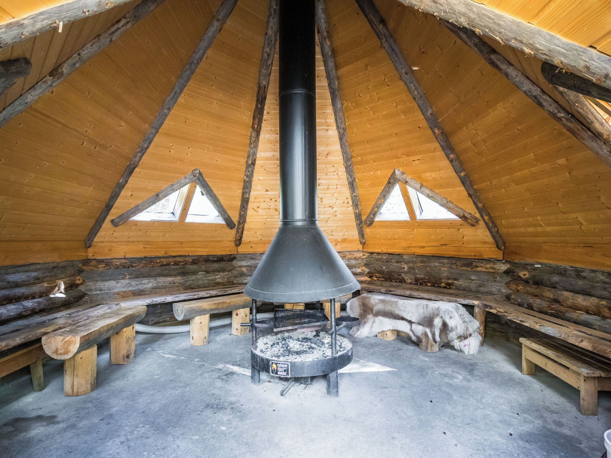 Une cabane en rondins avec un poêle à bois au centre, des bancs en bois et un chien.