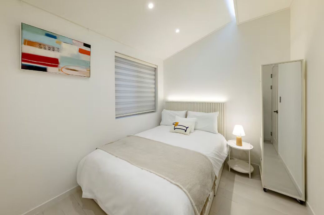 Habitación de hotel con cama, mesita de noche con lámpara, espejo y un cuadro en la pared.