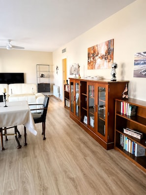 Ein Wohnzimmer mit Sofa, Esstisch, Bücherregal und Fernseher.