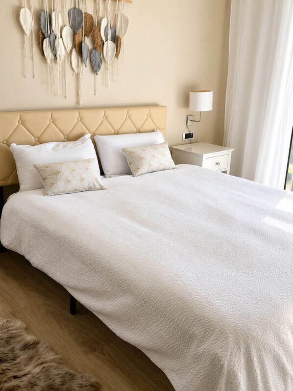 A bedroom with a tufted headboard, a large bed with white bedding, a nightstand with a lamp, and a wall decoration. - PARK de MAR Apartment (Sitges)