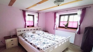 Ein Schlafzimmer mit Bett, Nachttischen, einem Fenster mit Vorhängen, einem Heizkörper und einer Deckenleuchte.