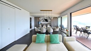 A modern interior with a dining area, a sofa set with patterned cushions, and a view through large windows. - Luxury Corba Lake View Apartment with Terrace (Canobbio)