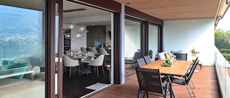 A modern outdoor dining area with a wooden deck, glass sliding doors leading to an interior dining space, and a view of the surrounding landscape.
