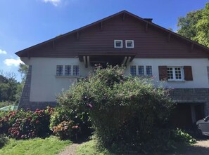 Una casa de dos pisos con techo marrón y paredes blancas, un porche delantero y un jardín con plantas florecientes.