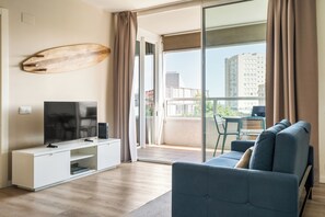 Een moderne woonkamer met een flatscreen televisie op een witte kast, een blauwe bank en een balkon met uitzicht op gebouwen.