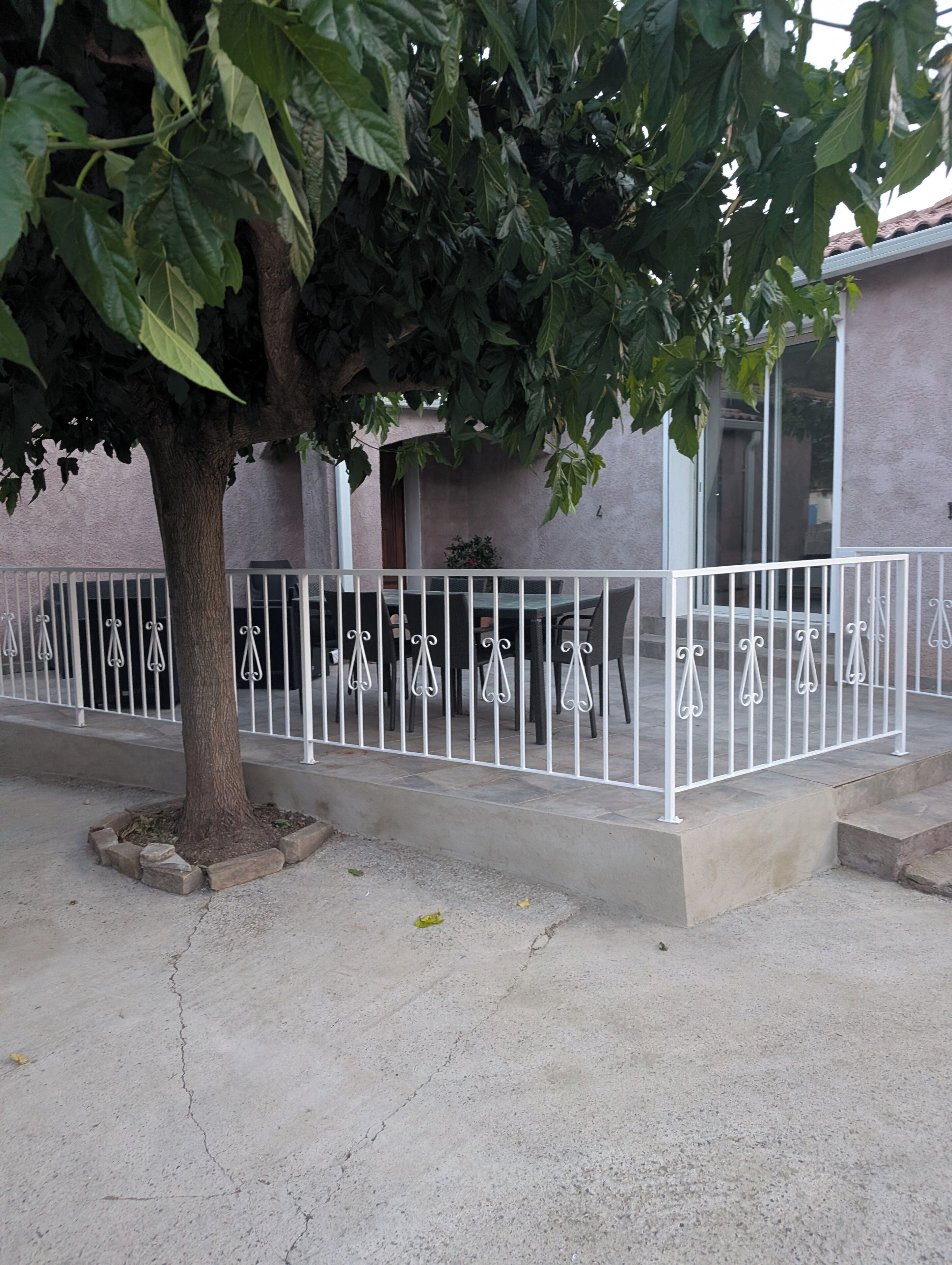 A tree-lined outdoor area with a white metal fence, a concrete floor, and a building with a window and door in the background.