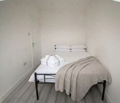 Una habitación pequeña con cama individual, colcha blanca, manta color beige y toallas blancas dobladas con cuidado.