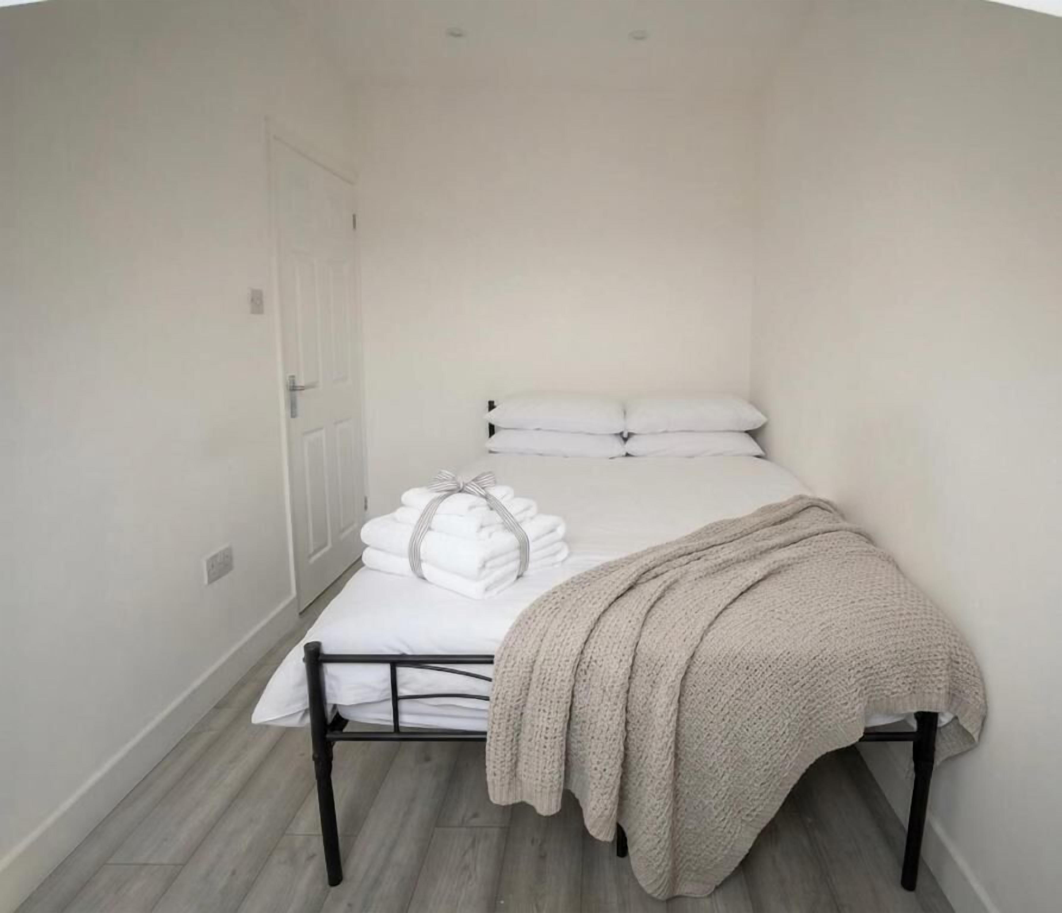 A small, single-bed room with a white door, a bed with white linens and a beige blanket, and a wooden floor.