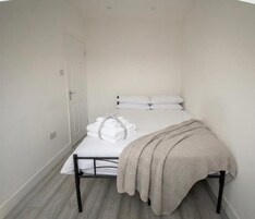 Una habitación pequeña con una cama individual, una puerta blanca, ropa de cama blanca y una manta color beige, y piso de madera.
