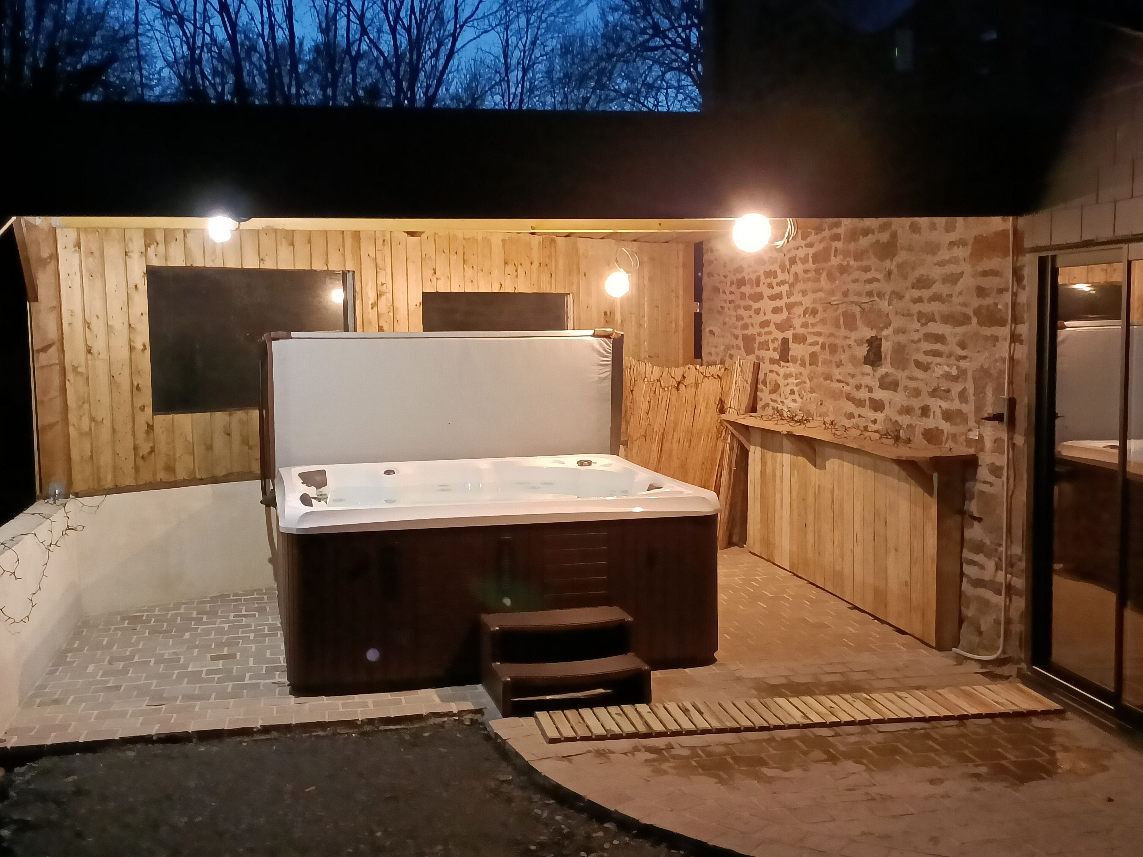 A hot tub in a wooden outdoor area with a stone wall and a glass door.