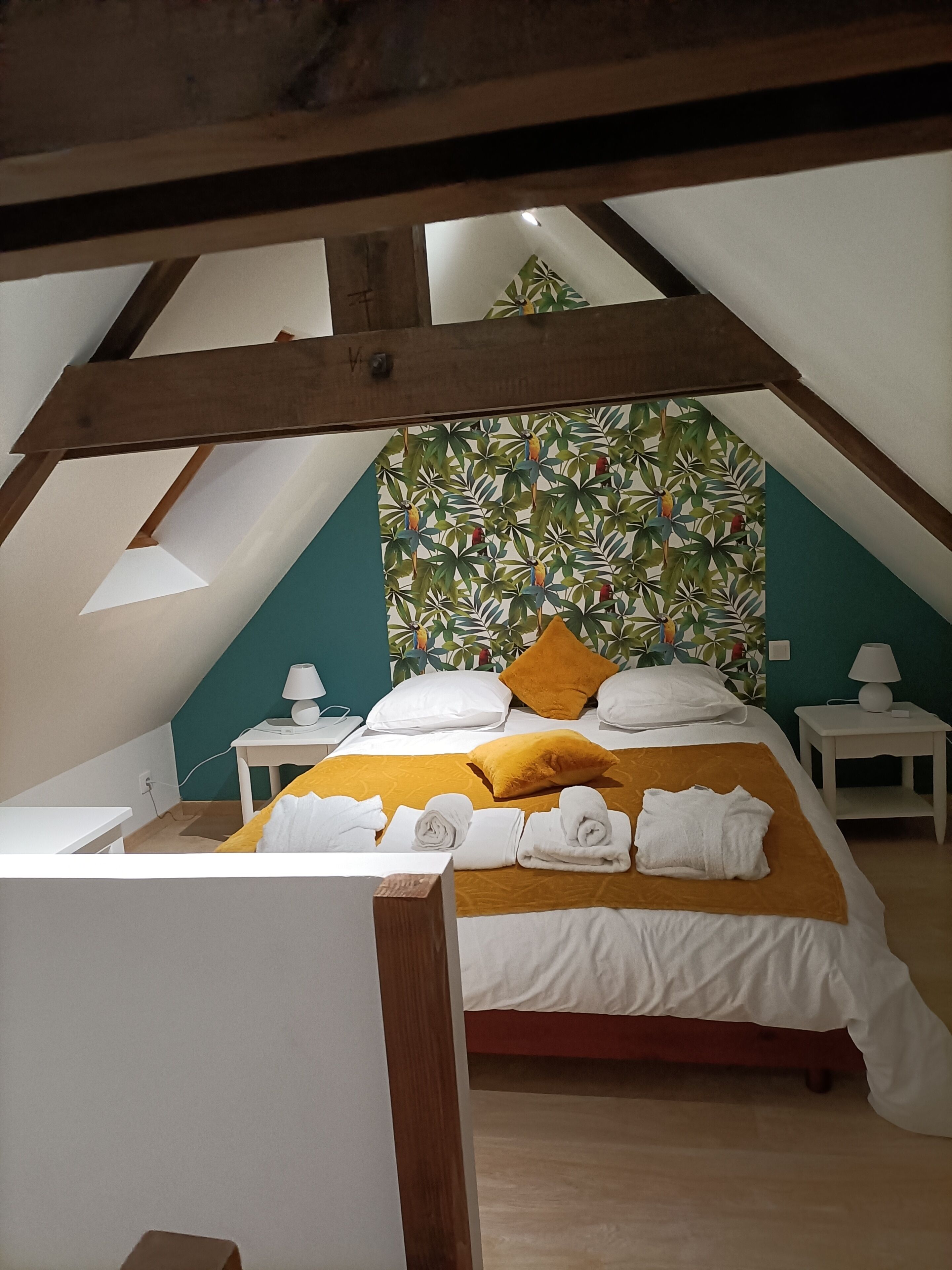 A bedroom with a sloped ceiling, a bed with white bedding and a yellow throw, a wall with a leaf pattern, and a small bedside table with a lamp.