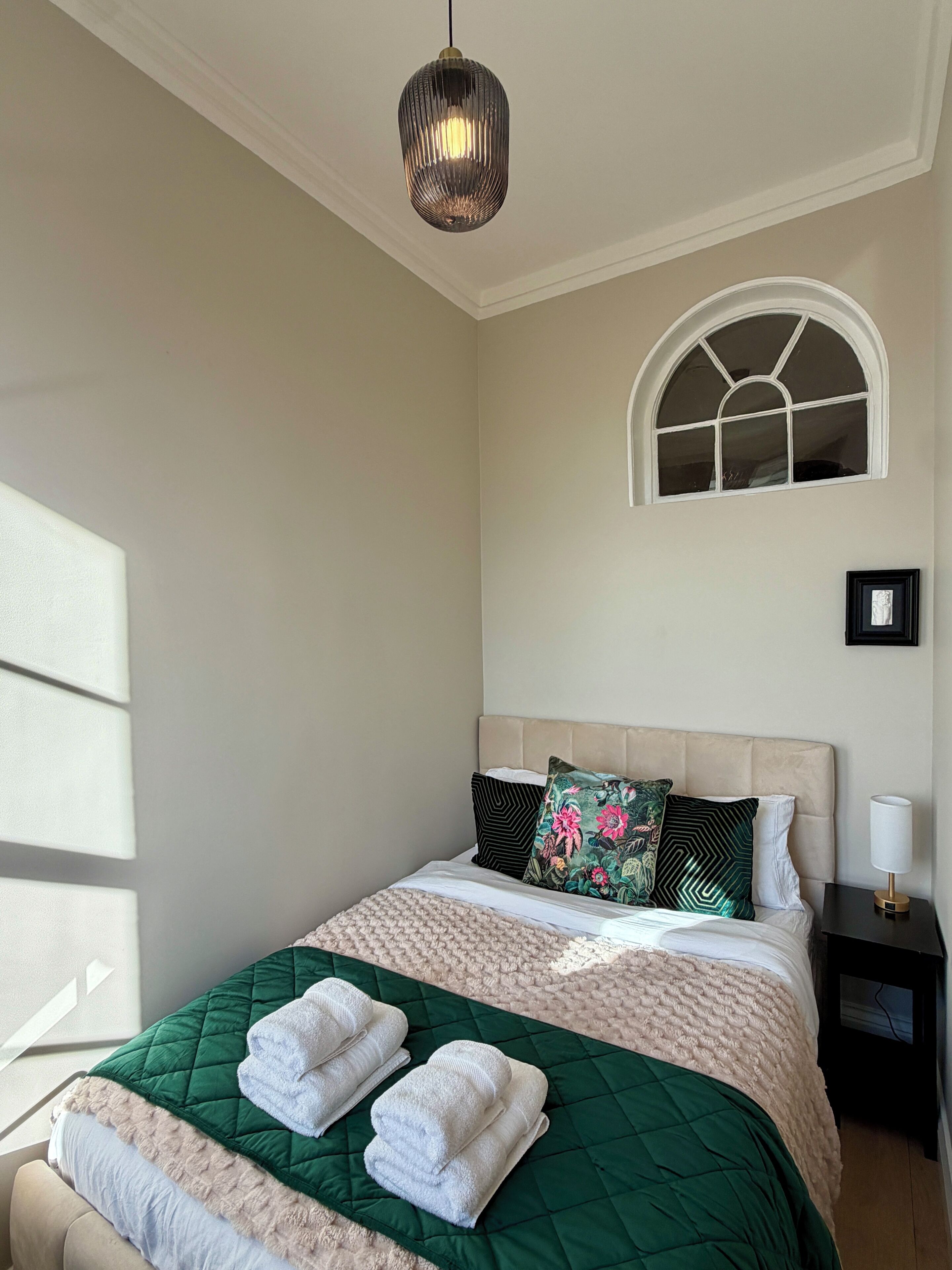 A bedroom with a bed, pillows, a bedside table, a lamp, a window, and a hanging light fixture.