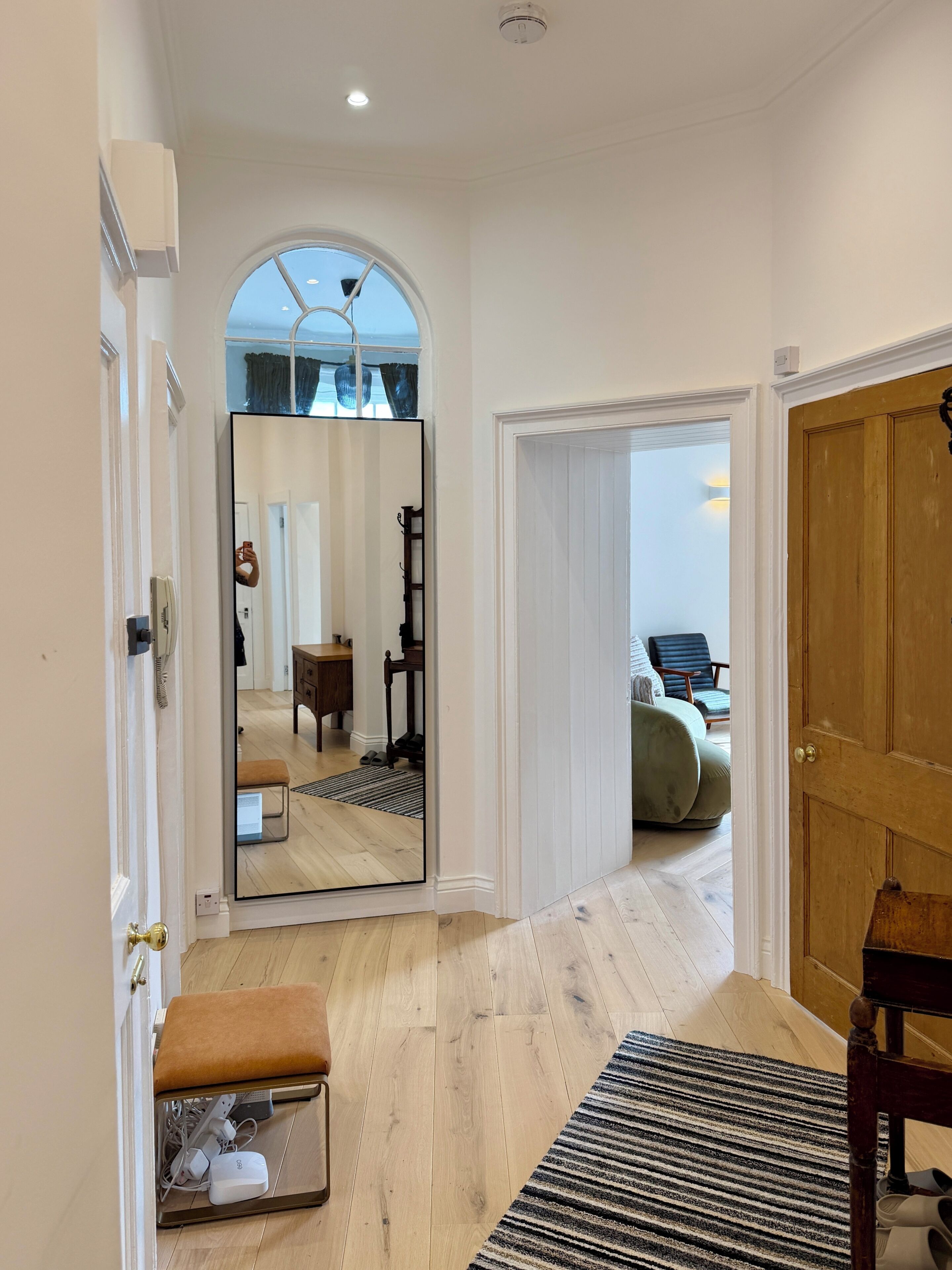 A hallway with a large mirror reflecting a room, a wooden bench, a rug, and a door leading to another room.
