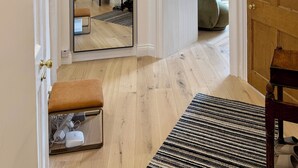 Een gang met een grote spiegel die een kamer weerspiegelt, een houten bankje, een vloerkleed en een deur naar een andere kamer.