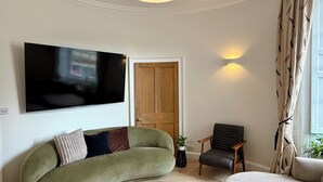 Un saló amb un sofà corbat, un televisor de pantalla plana, una porta de fusta, una taula petita amb una planta i una finestra amb cortines.