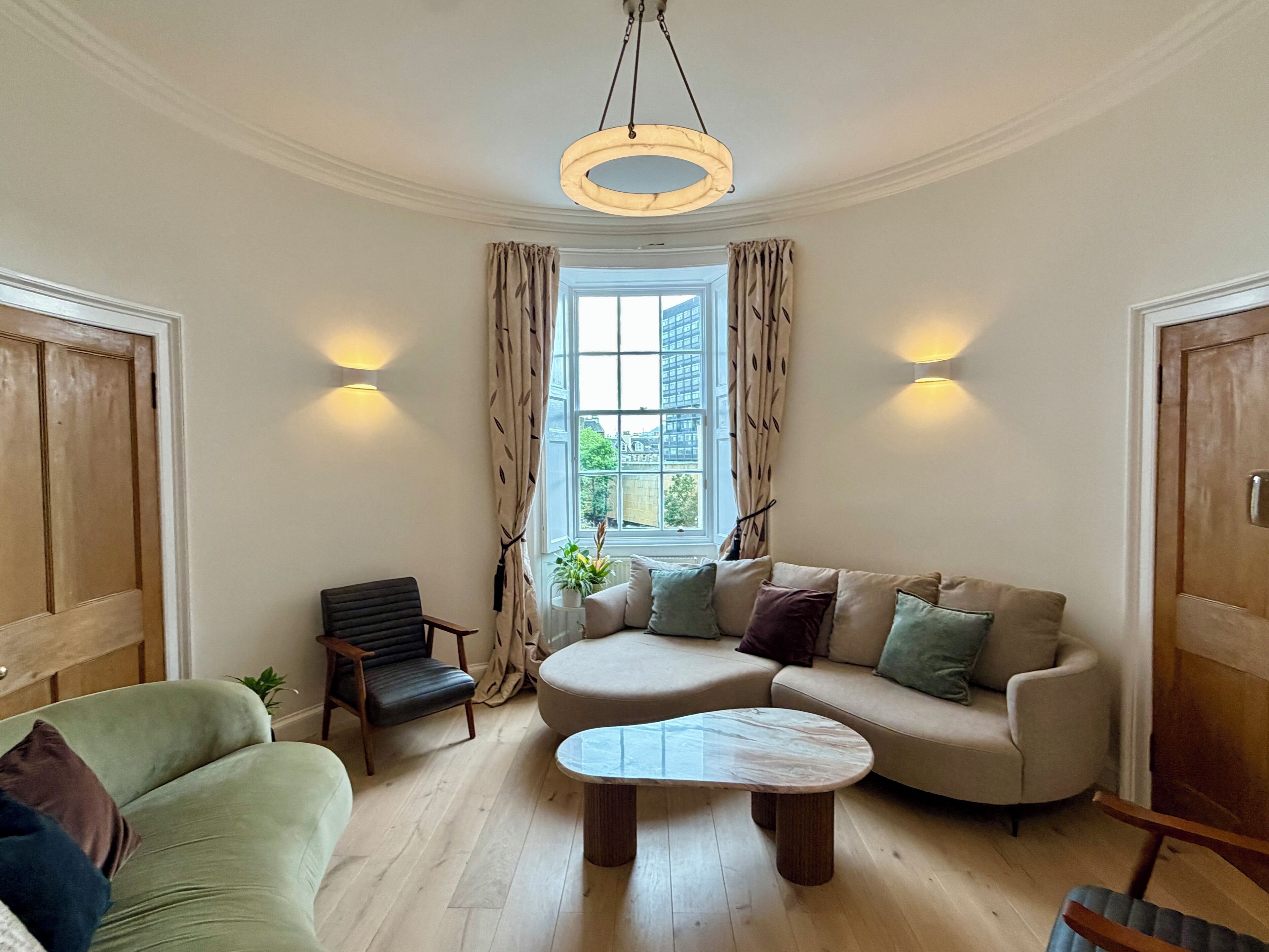 A living room with a sofa, armchair, and coffee table. There is a window with curtains and a view of buildings outside.