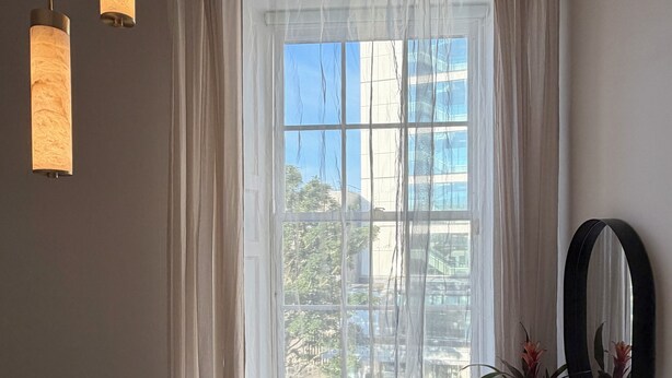 Un dormitori amb un llit, una petita arca, un mirall i una finestra amb cortines transparents.