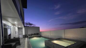 A rooftop pool area with a hot tub and seating, surrounded by a low wall and a clear sky.