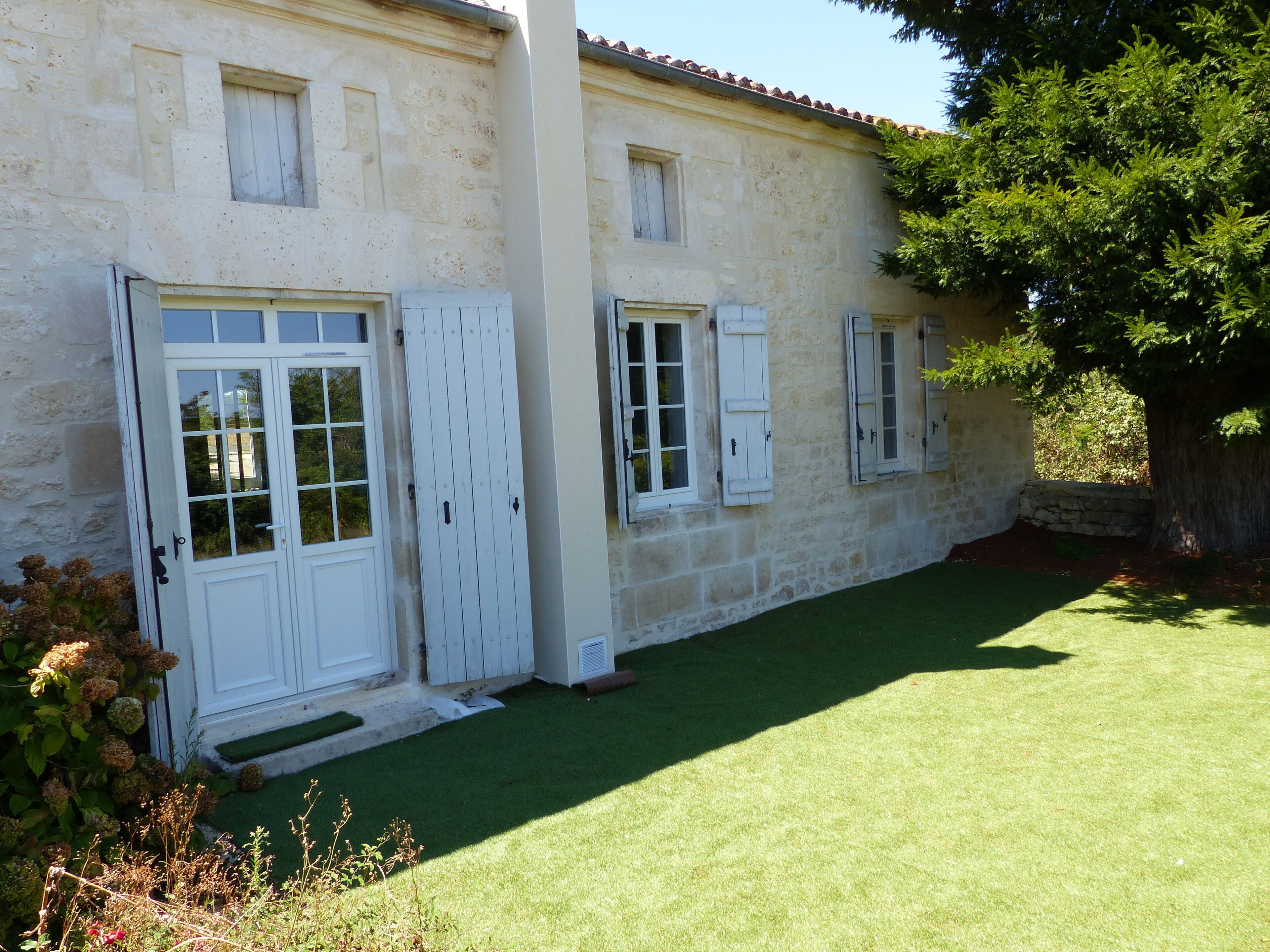 A stone building with white doors and shutters, a well-maintained lawn, and a tree to the right.