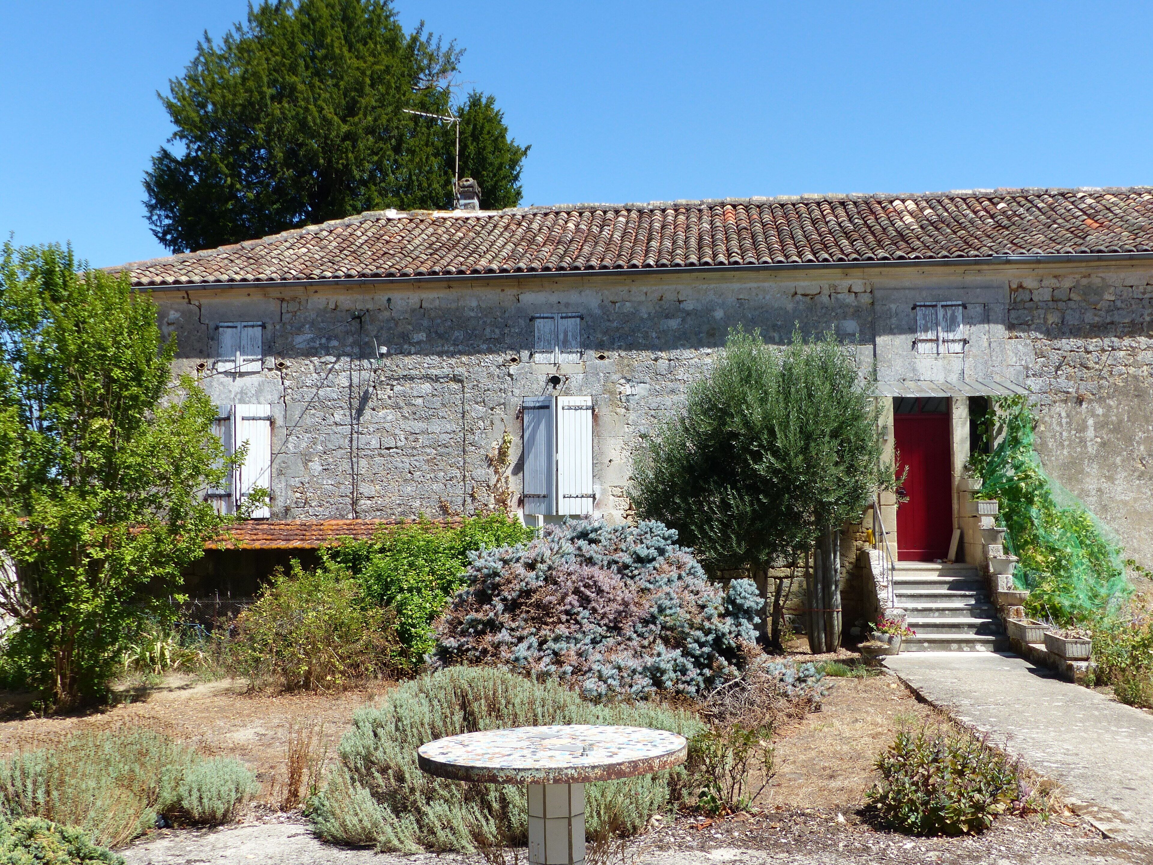 A rustic stone building with a tiled roof, a red door, and a small garden with a round table.