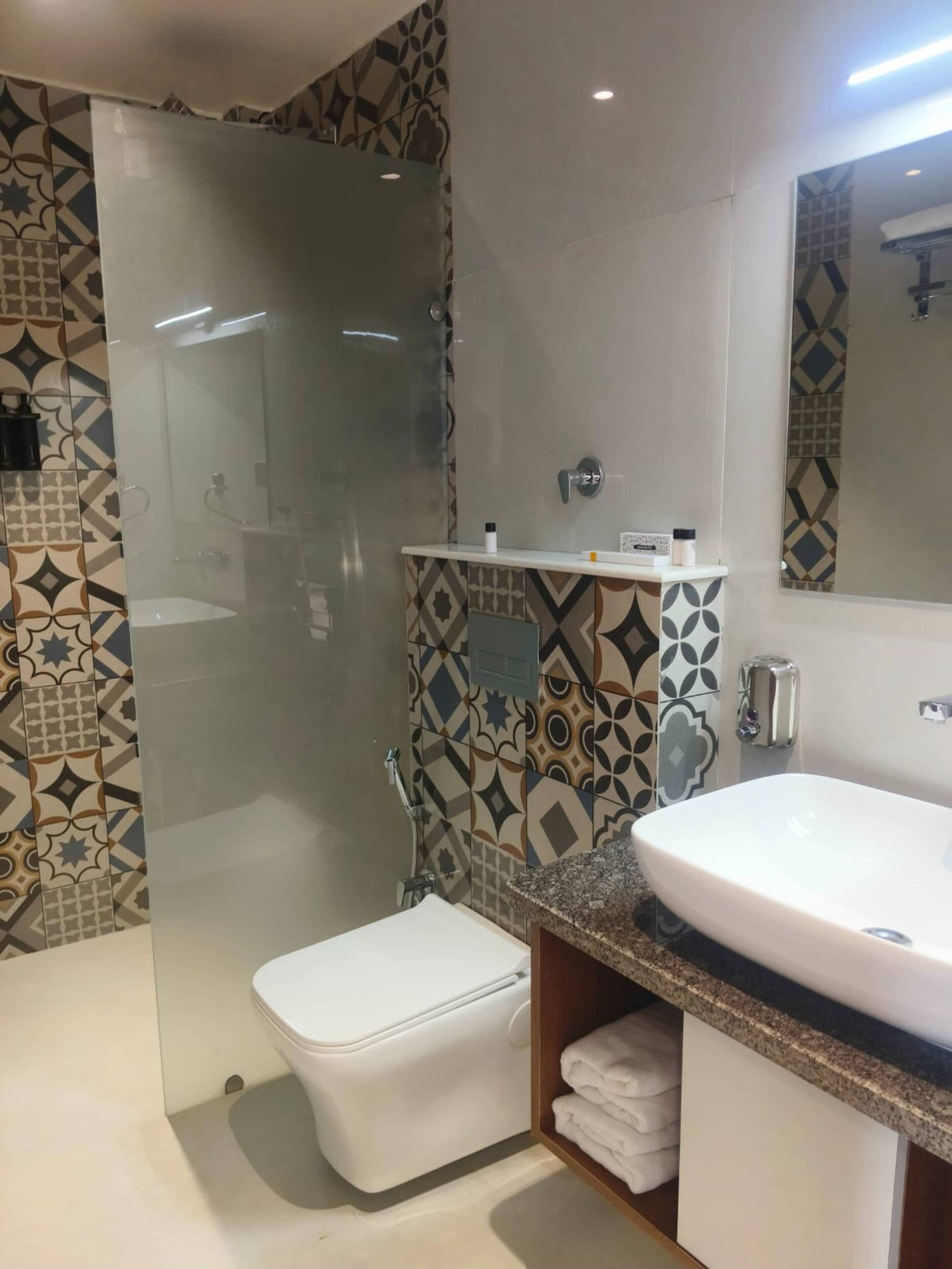Une salle de bain dotée d’une douche à parois de verre, de toilettes blanches, d’un lavabo avec un plan de travail en granit et de carreaux décoratifs sur les murs.
