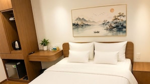 Habitación de hotel con cama, mesita de noche, un armario pequeño, un cuadro y un refrigerador.