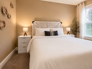 A bedroom with a bed, two bedside tables, a nightstand, a wall-mounted lamp, and a window with curtains. - In-ground Pool | 5BR | Family Fun (Warner Robins)