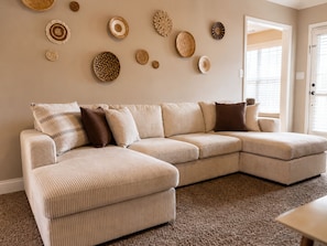 A beige sectional sofa with a textured fabric, a brown and beige striped throw pillow, and a small round table with a light-colored surface. - In-ground Pool | 5BR | Family Fun (Warner Robins)