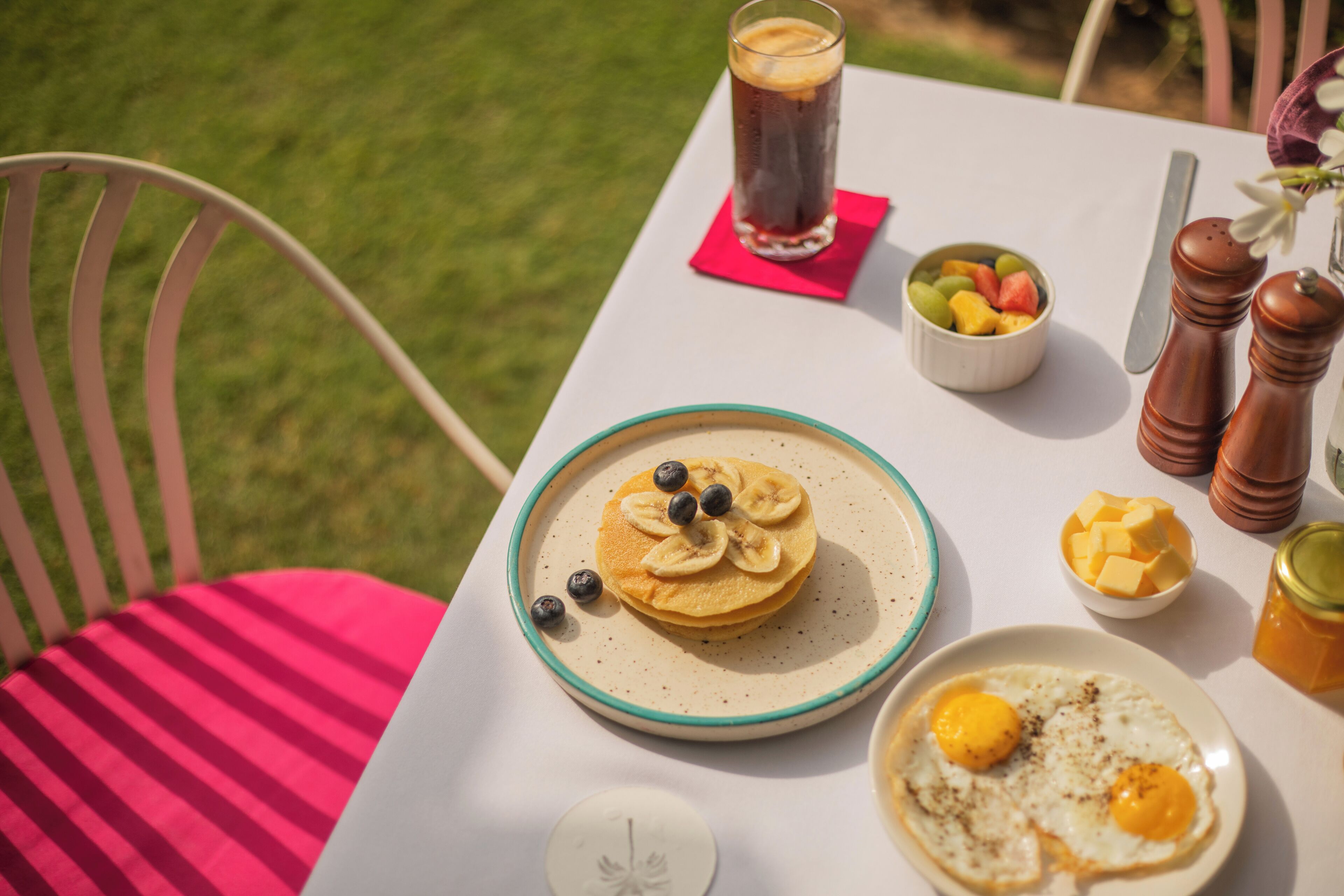 Una mesa de desayuno con panqueques, un vaso de café helado, un bol de fruta y un plato con un huevo.