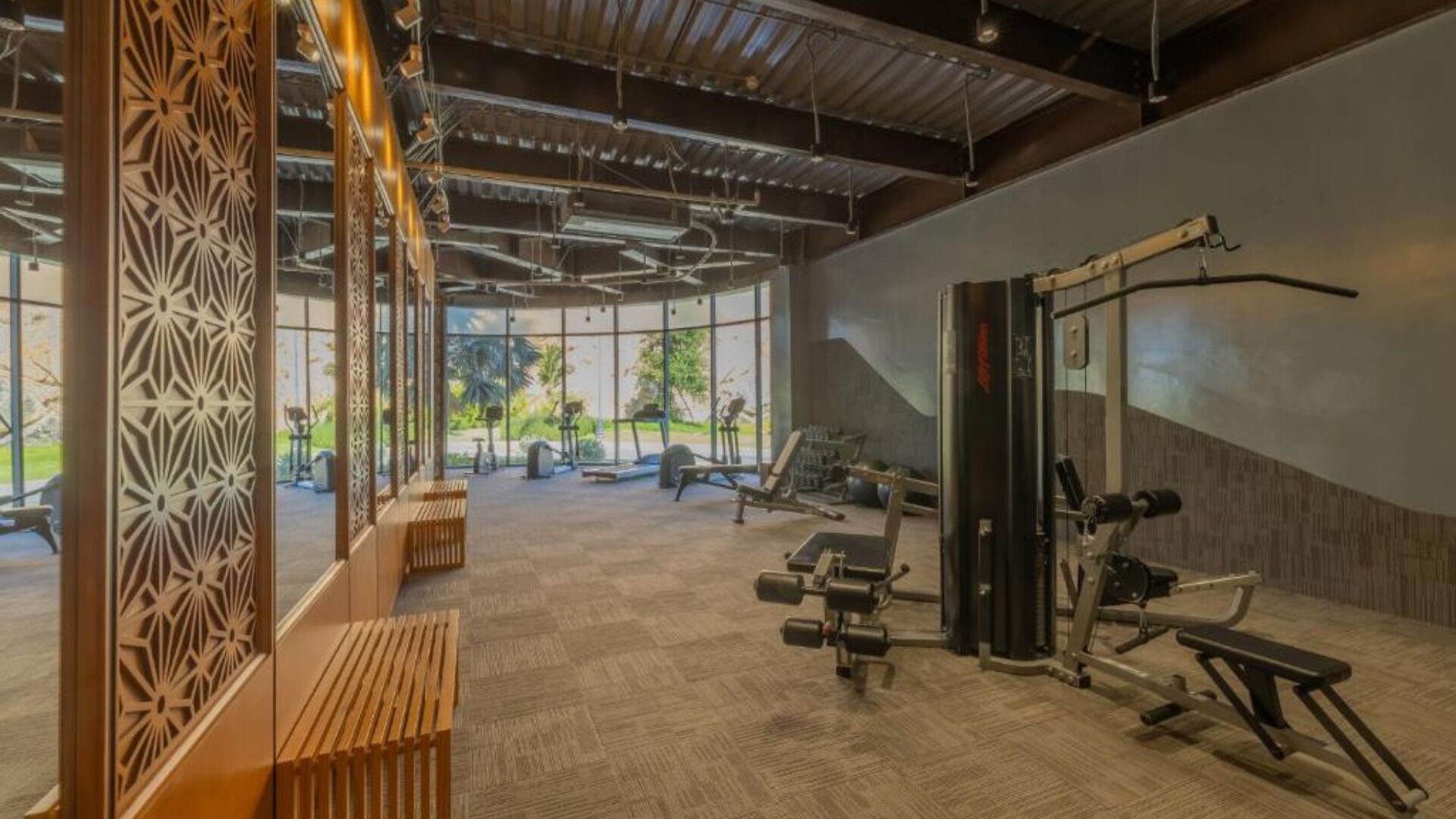 A gym with exercise equipment, a wooden bench, and large windows offering a view of greenery.