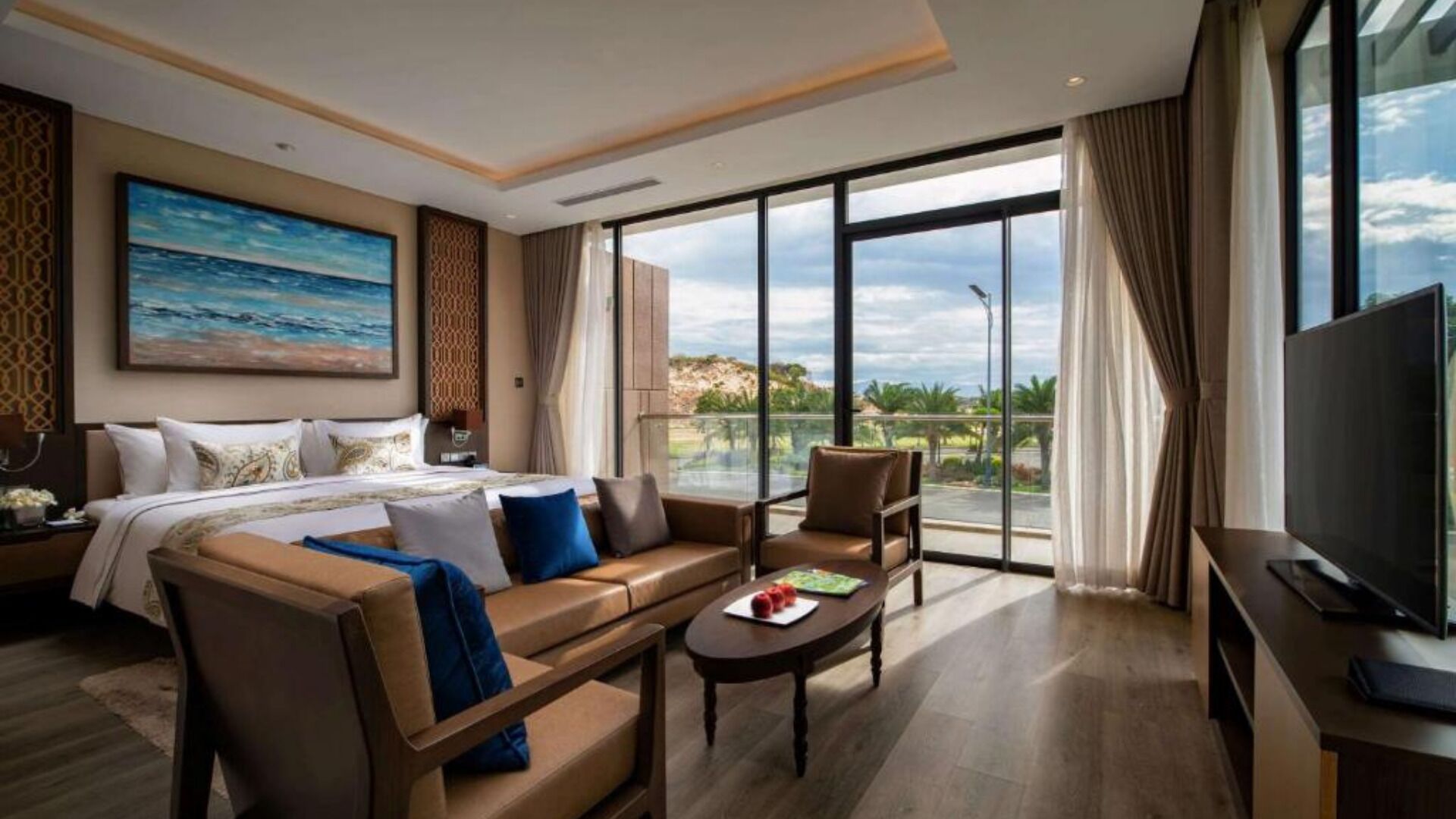 A hotel room with a bed, a sofa, a coffee table, a TV, and a balcony with a view.