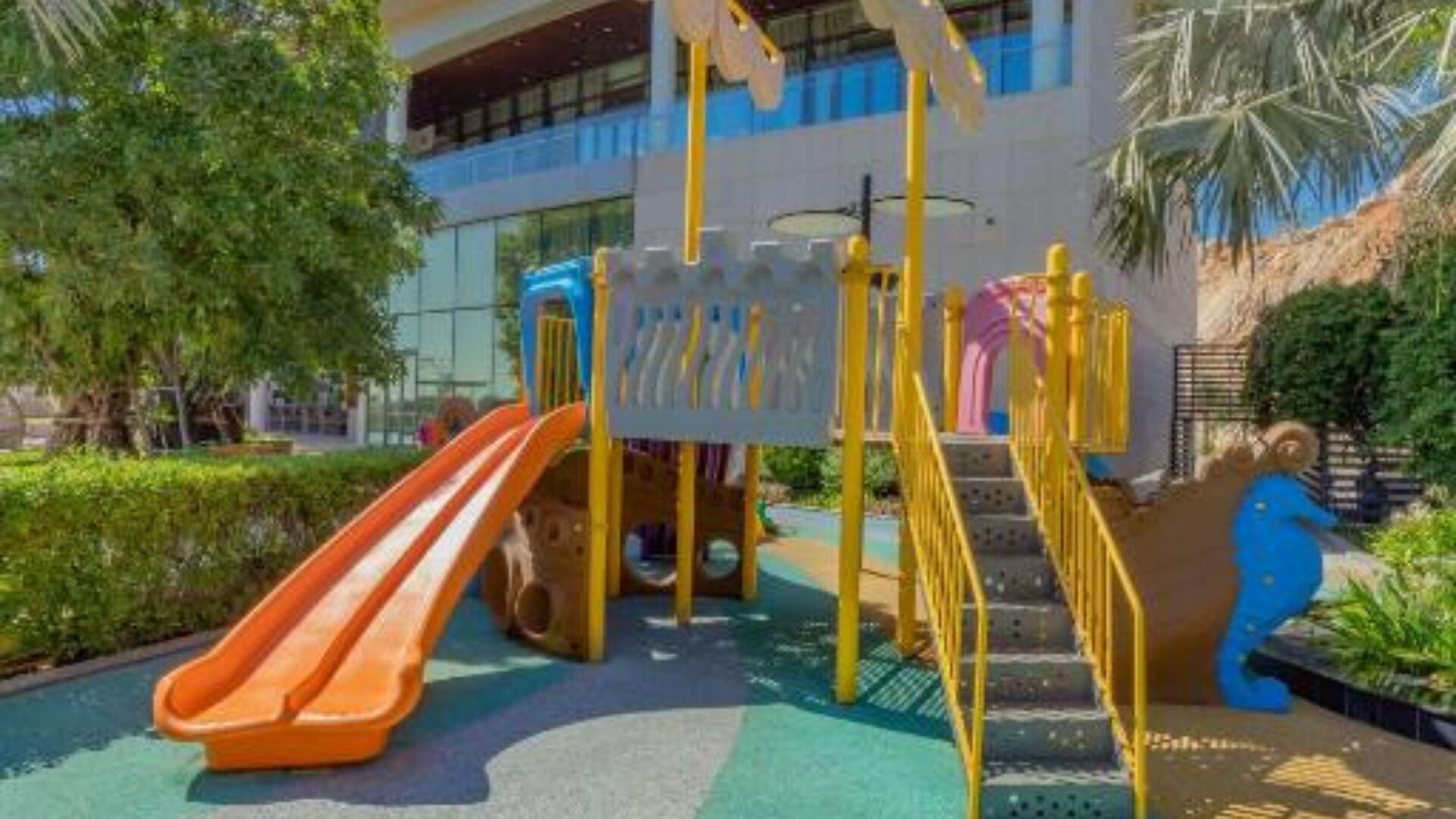 Un parque infantil con tobogán y estructuras para escalar, rodeado de vegetación y un edificio al fondo.