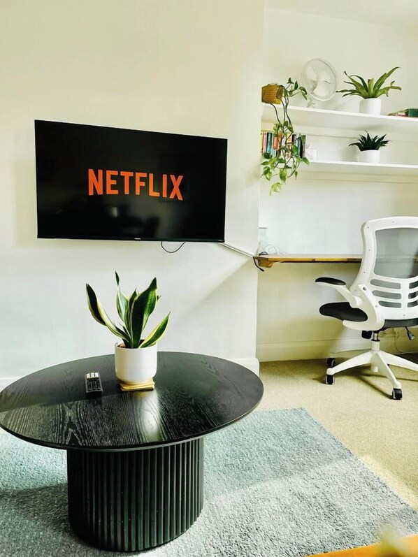 A modern living room with a round black coffee table, a potted plant, a white office chair, a mounted flat-screen TV, and a bookshelf with books and decorative items.