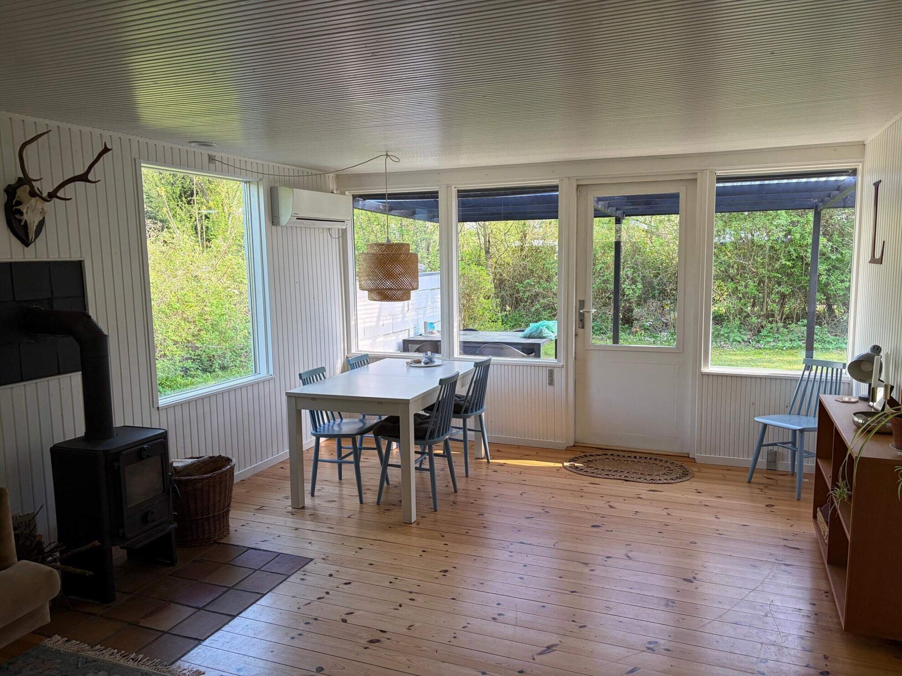 A room with a wooden floor, a table with chairs, a fireplace, and large windows.