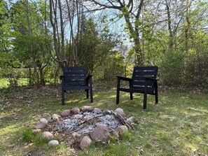 Two black chairs facing a fire pit surrounded by rocks in a wooded area.