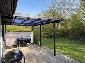 Eine überdachte Terrasse mit Grill, Gartenmöbeln und Blick auf einen Garten.