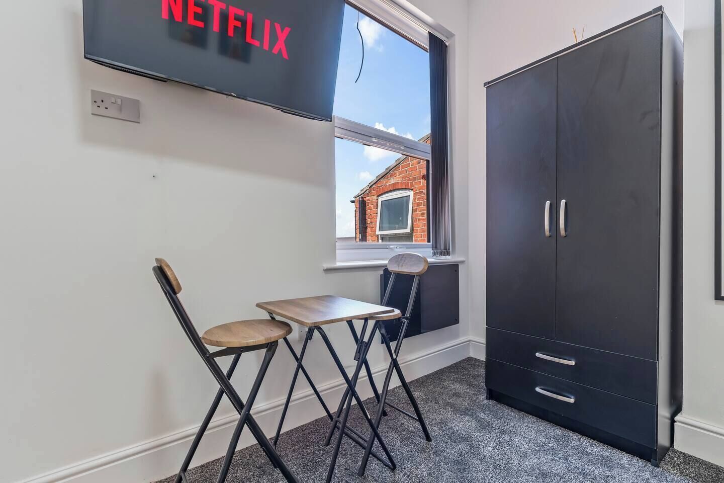 Een kamer met een televisie waarop 'NETFLIX' staat, een raam met uitzicht op een bakstenen gebouw, een zwarte garderobe en een kleine tafel met twee stoelen.