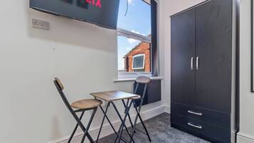 Een kamer met een televisie waarop 'NETFLIX' staat, een raam met uitzicht op een bakstenen gebouw, een zwarte garderobe en een kleine tafel met twee stoelen.
