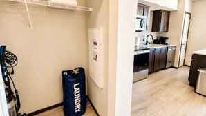 A laundry bag labeled 'LAUNDRY' in a hallway with a white wall, a shelf with a plant and towels, and a glimpse of a modern kitchen with dark cabinets and a white countertop. - Stylish 2BR Tacoma Stay | Near Hospitals & Tacoma Dome (Tacoma)