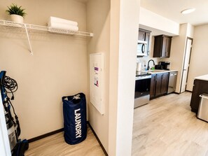 En tvättkorg märkt med texten ”TVÄTT” står i en hall med en vit vägg, en hylla med en växt och handdukar, och man kan skymta ett modernt kök med mörka skåp och en vit bänkskiva.