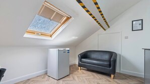 Ein Zimmer mit schrägem Dach, Deckenventilator, einem weißen Kühlschrank, einer schwarzen Ledersofa und einem Holzboden im Fischgrätmuster.