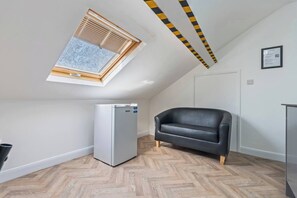 A room with a sloped ceiling, a built-in ceiling fan, a white refrigerator, a black leather sofa, and a wooden herringbone floor. - Top Floor Studio Apartment with Skylight & Fast WiFi in Ilkeston (Derbyshire)