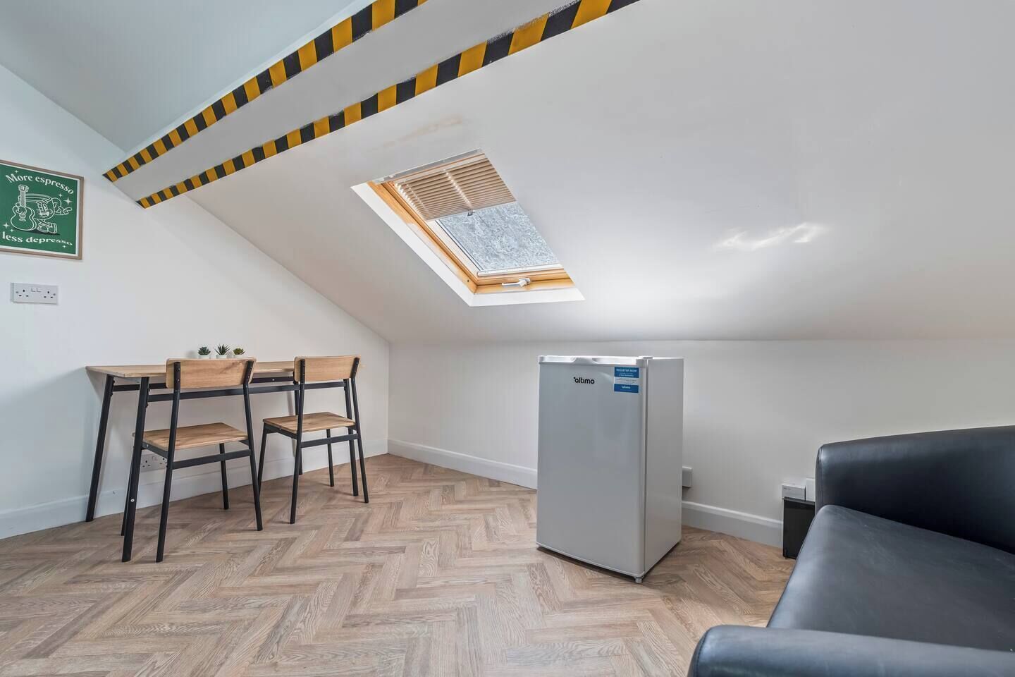Una habitación pequeña y moderna con suelo de madera en patrón chevron, un sofá negro, un refrigerador blanco, una mesa de madera con sillas y un tragaluz.